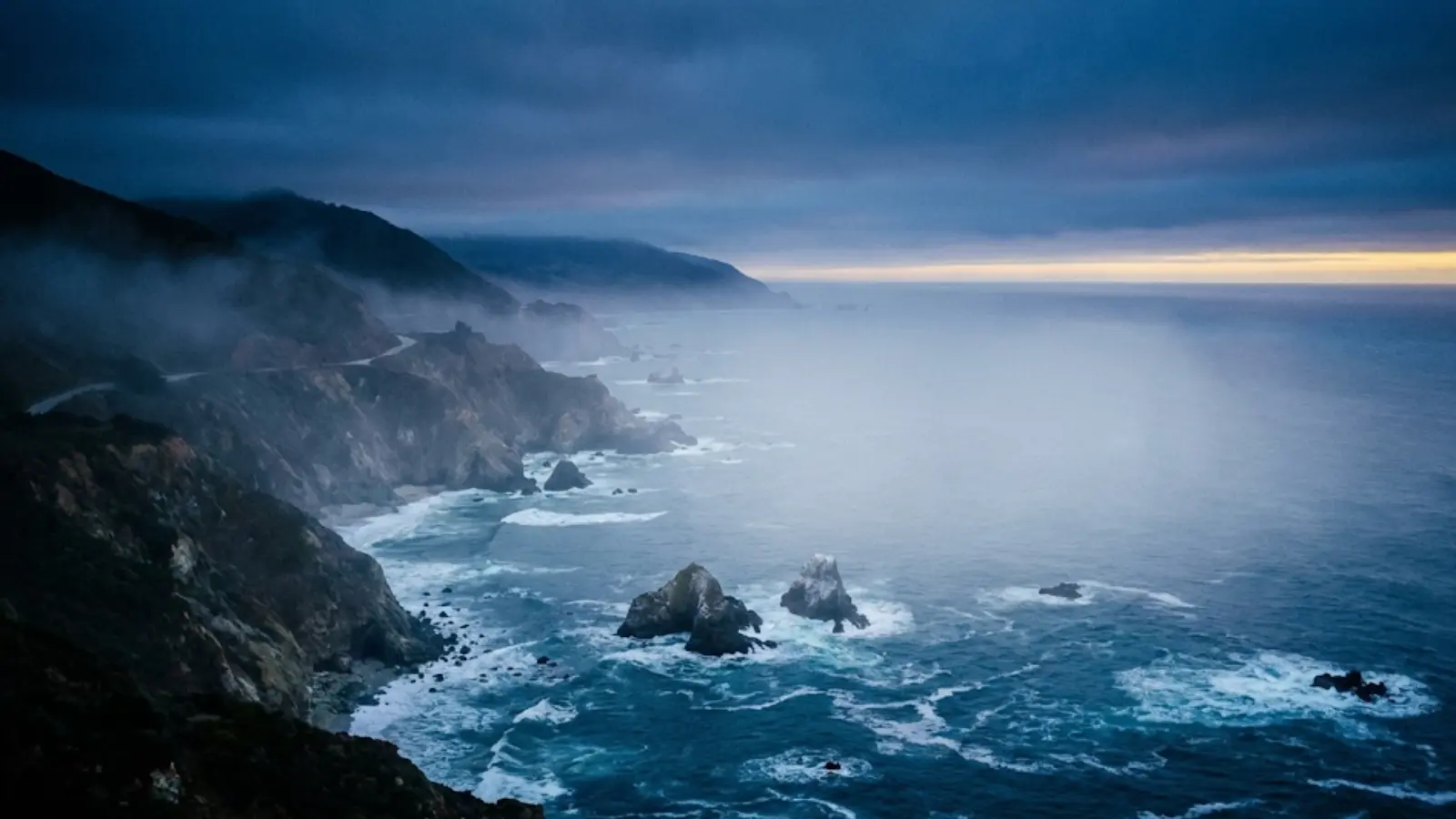 Dramatic California coastline at Big Sur during blue hour with misty cliffs and deep ocean waves.